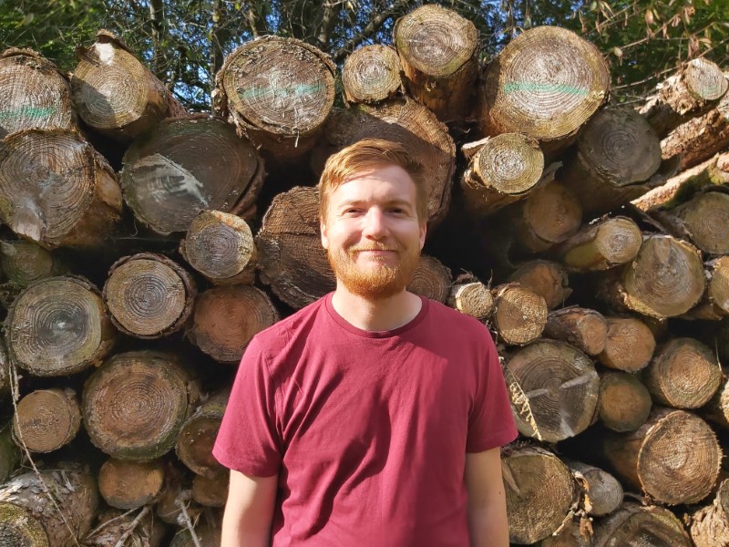 Me with a beard in front of loads of logs